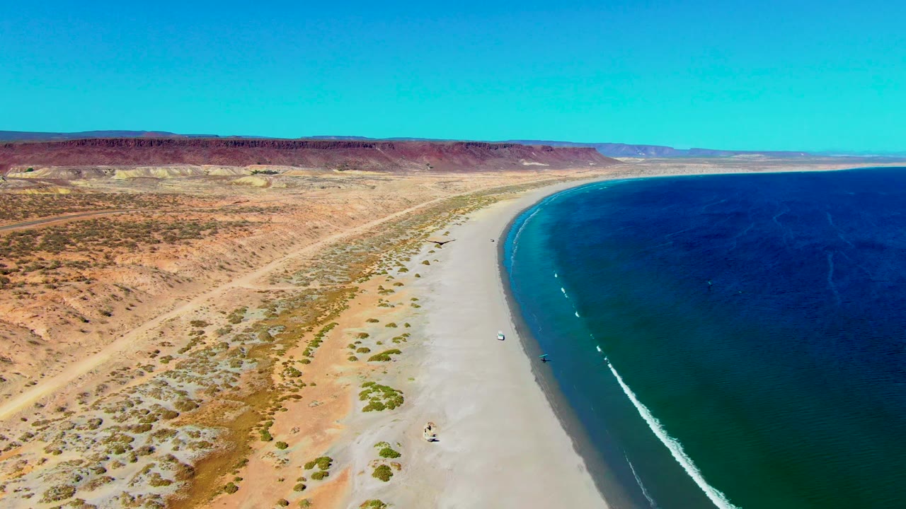 Aerial San Juanico coastline frame with a bright white wave line near the beach, clear blue water to the right, arid land to the left, and no visible people.