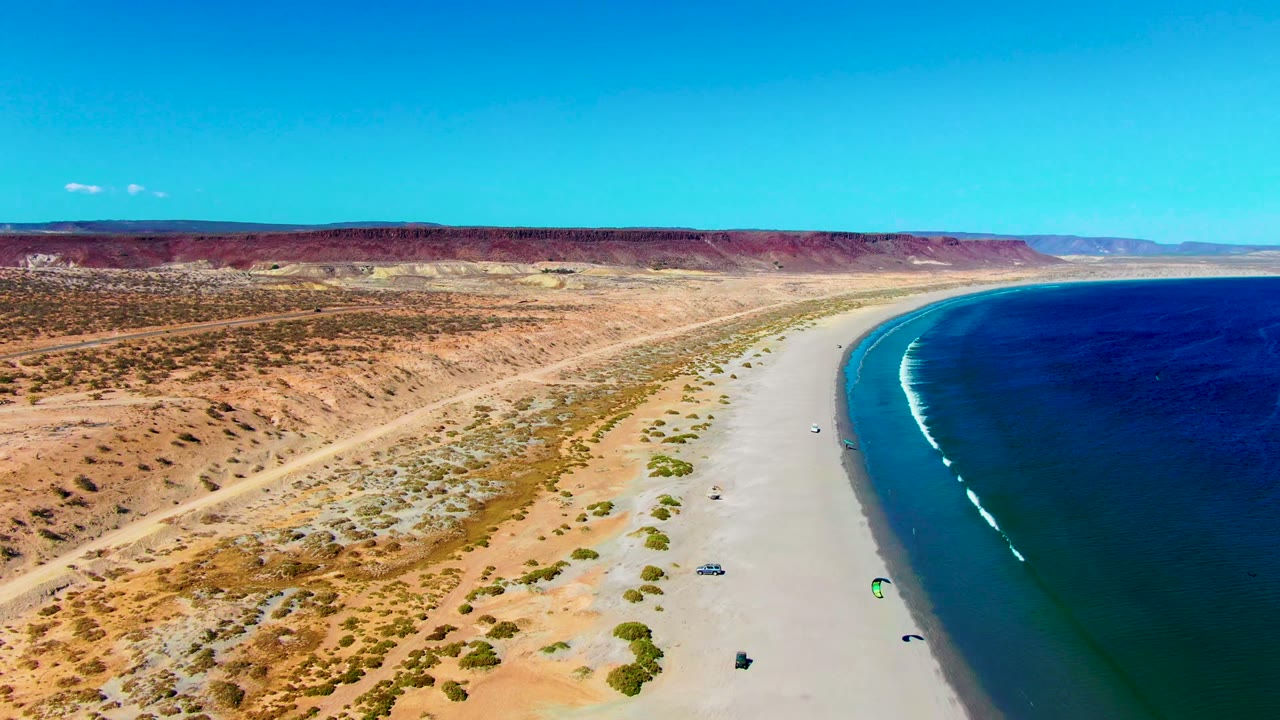 Aerial frame from San Juanico, Baja California Sur showing a wide sandy beach, deep blue ocean on the right, desert scrub and road on the left, and tiny kite surfers near the shoreline.