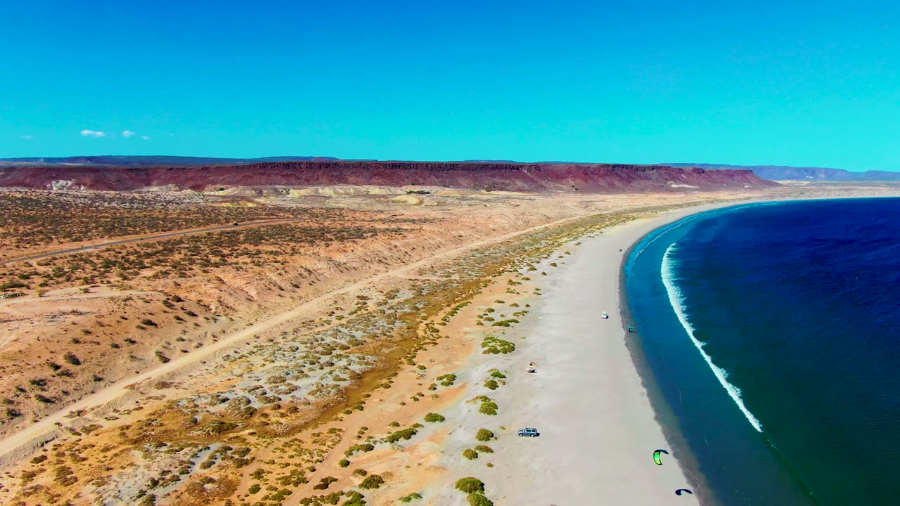 Aerial view over San Juanico with the dirt road and dry scrub more prominent at left, a pale beach through the center, tiny kite surfers near the waterline, and rolling ocean waves at right.
