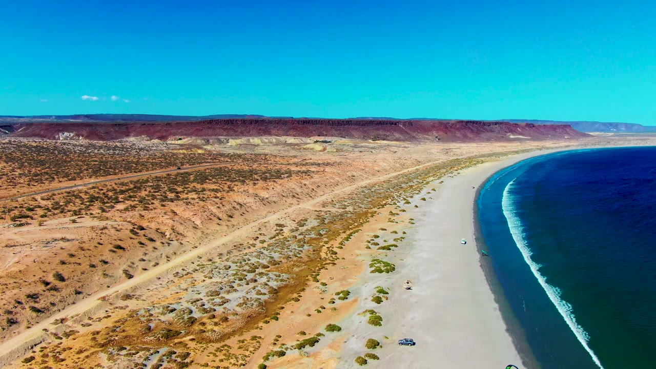 San Juanico aerial frame with the coastline curving along the right edge, open desert and scrub filling the left side, tiny kite surfers below, and low mesas under clear blue sky.