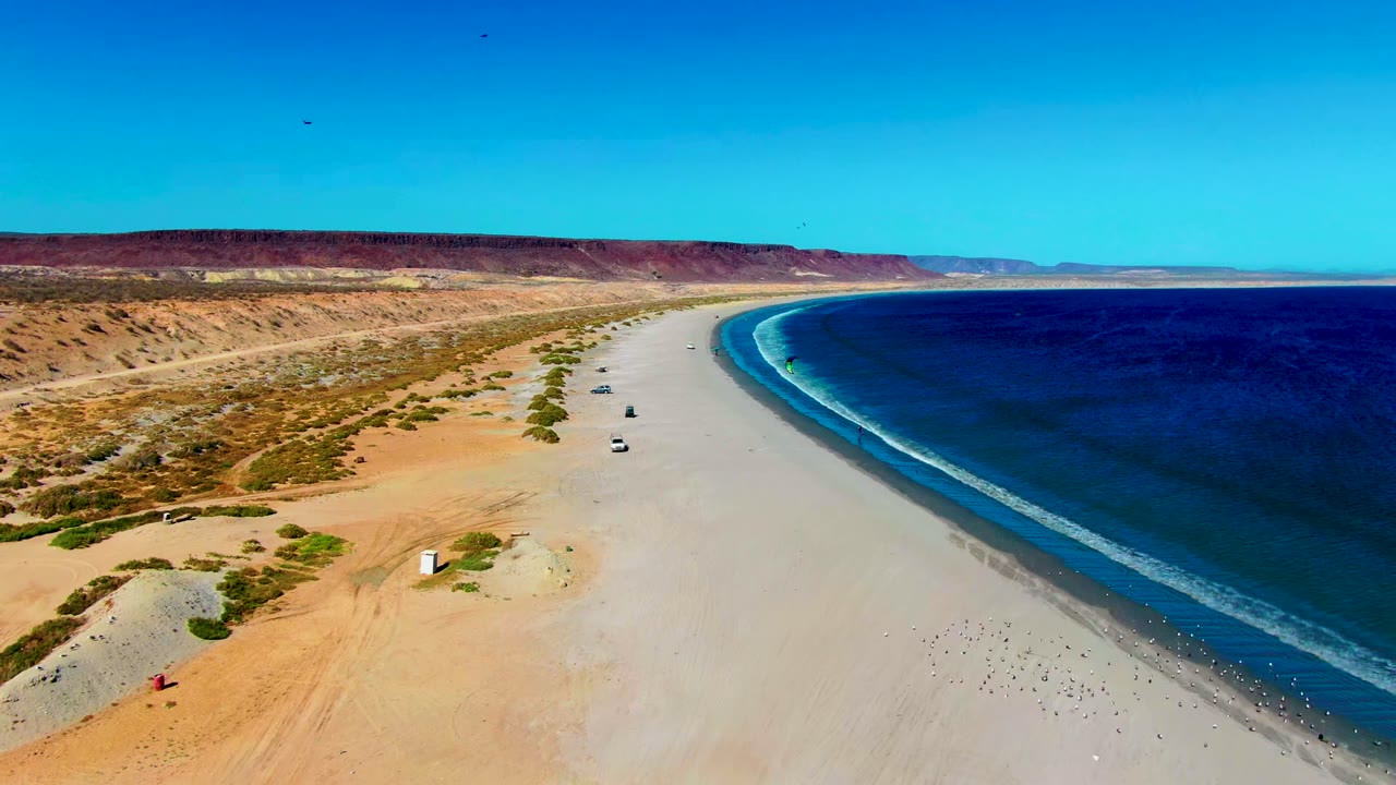 Aerial frame over San Juanico, Baja California Sur, showing a wide pale beach, deep blue ocean, desert dunes, small vehicles on the sand, and no visible people.