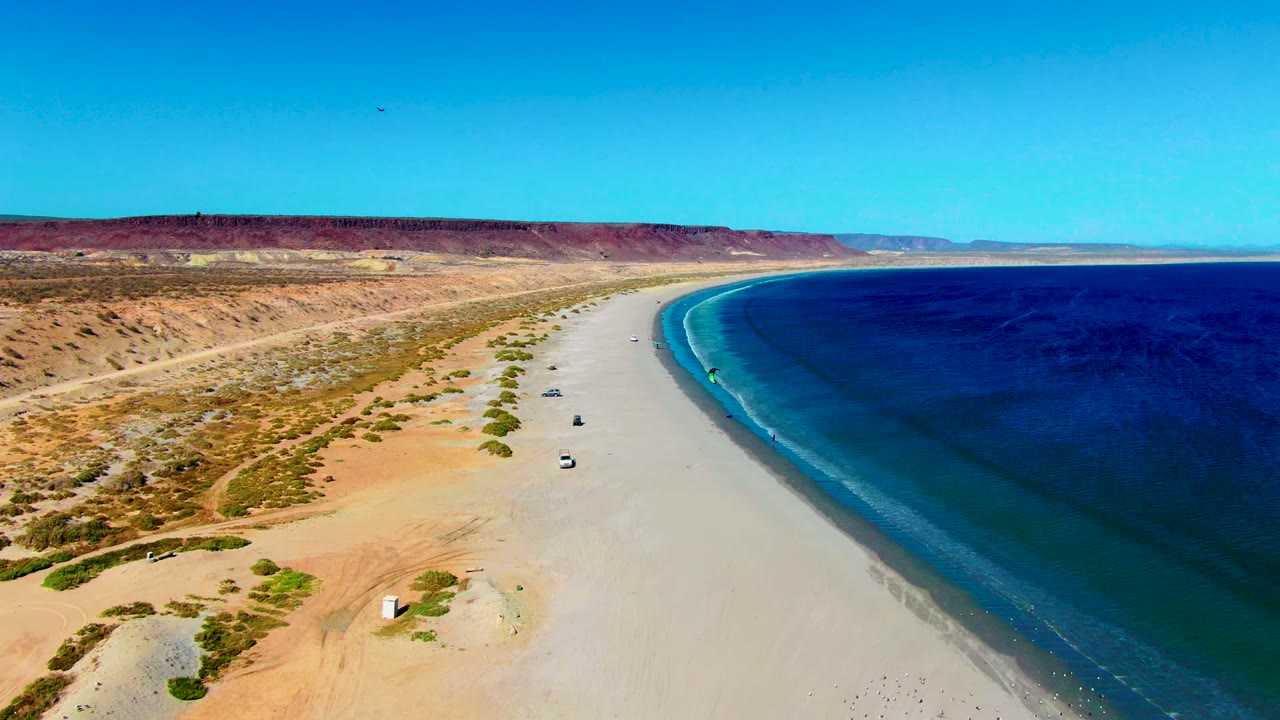The San Juanico coastline fills the frame with desert dunes to the left, small vehicles on the pale beach, dark blue water to the right, and no visible people.
