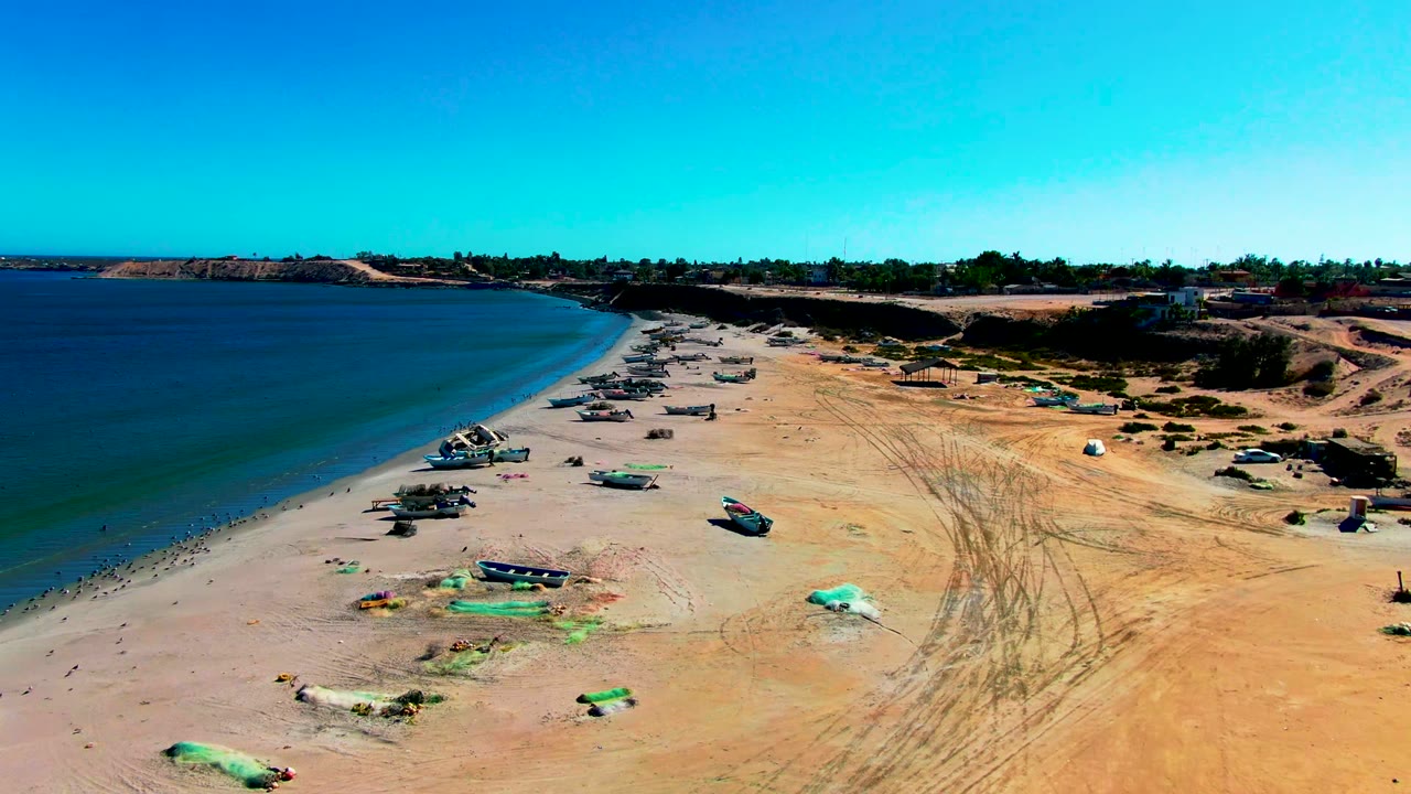 Aerial view in San Juanico, Baja California Sur, with small fishing boats scattered on sand beside calm blue water and no visible people.