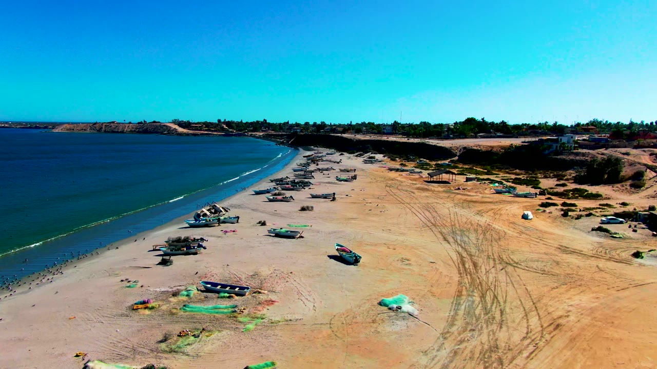 A wide aerial frame of San Juanico beach shows small boats on pale sand, a narrow white wave line, scrub beyond the beach, and no visible people.