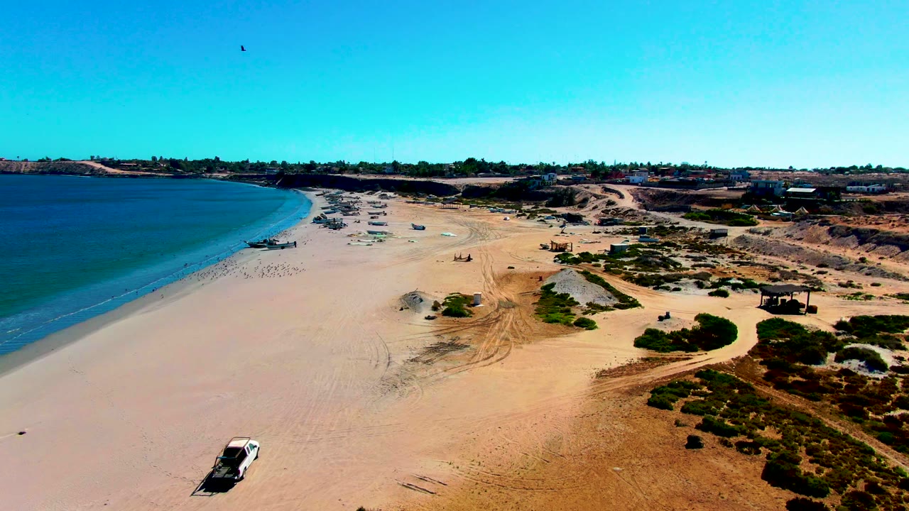 Aerial frame over San Juanico, Baja California Sur, with blue ocean on the left, a wide sandy beach, dune tracks, distant beach activity, and a white car near the lower left edge.