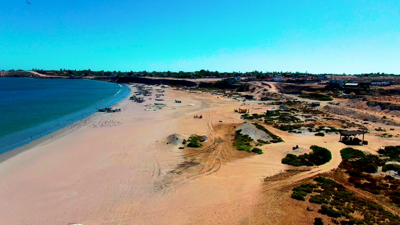 Wide aerial beach view with ocean, dunes, tire tracks, and scattered objects along the shore.