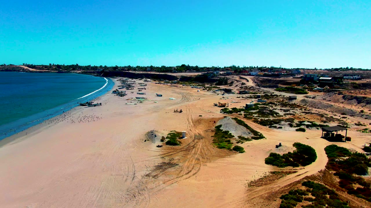 San Juanico aerial frame with a curved blue shoreline on the left, open tan beach across the center, scattered small figures and vehicles near the surf, and dunes rising toward distant buildings.