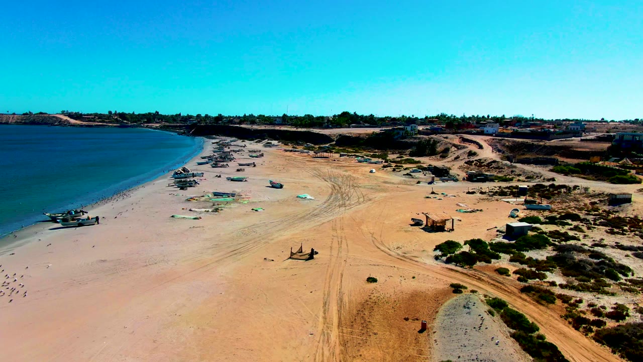 Aerial frame over San Juanico, Baja California Sur showing beached fishing boats, green tarps, tire tracks, blue water, dunes, and no visible people.