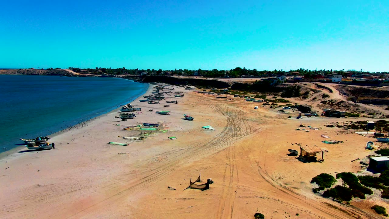 Aerial view in San Juanico, Baja California Sur with beached boats set farther along the left shoreline, green tarps on pale sand, strong blue sky, and no visible people.