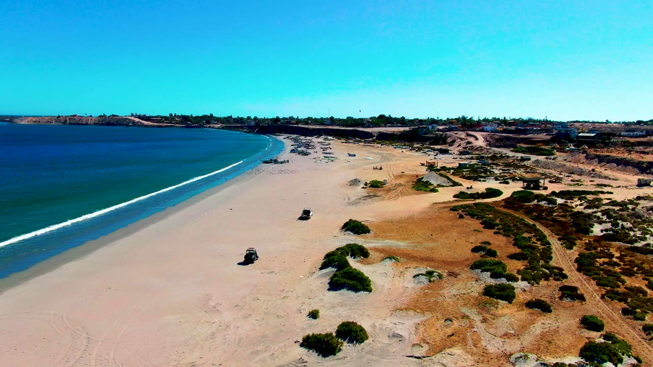Aerial frame from San Juanico, Baja California Sur showing a dark 4x4 on pale beach sand with blue ocean to the left, scrub-covered dunes to the right, and no visible people.