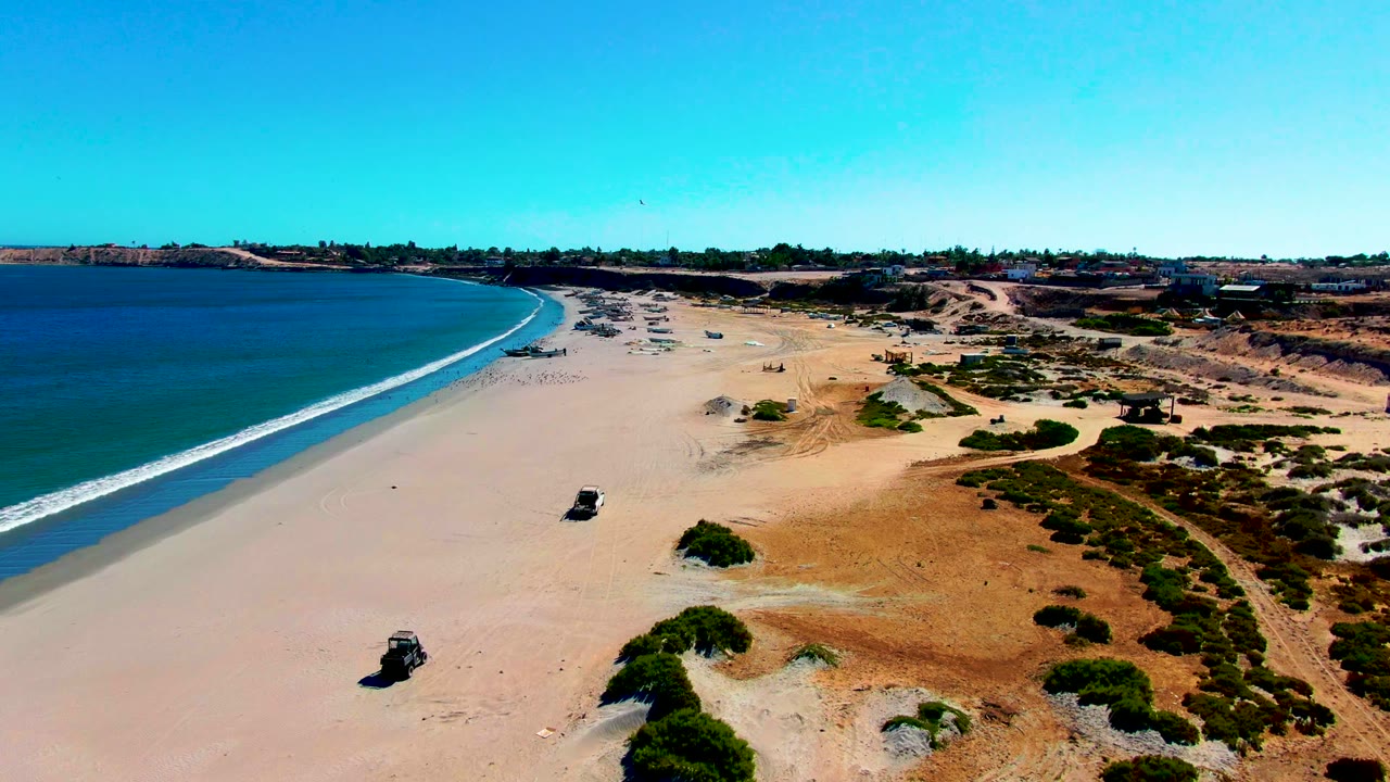 San Juanico aerial frame with the 4x4 slightly closer in the lower beach area, blue water and a white surf edge at left, dry dune vegetation at right, and no visible people.