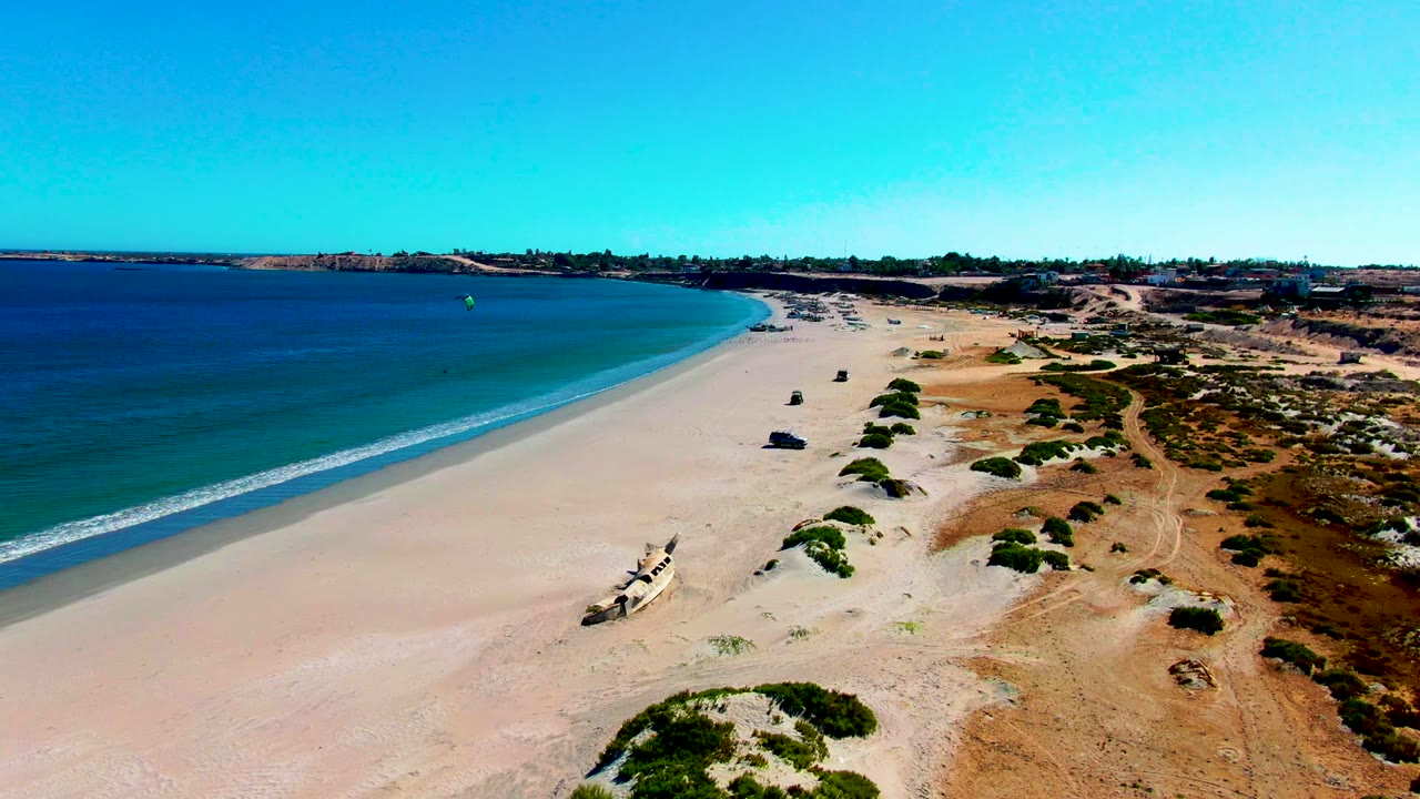 Aerial frame over San Juanico beach with blue ocean on the left, pale sand in the center, dry dunes and scattered shrubs on the right, and no visible people.