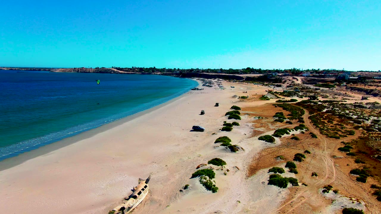 Aerial shoreline frame over San Juanico with turquoise water, a clean curve of open sand, patchy dune vegetation, dry terrain beyond the beach, and no visible people.