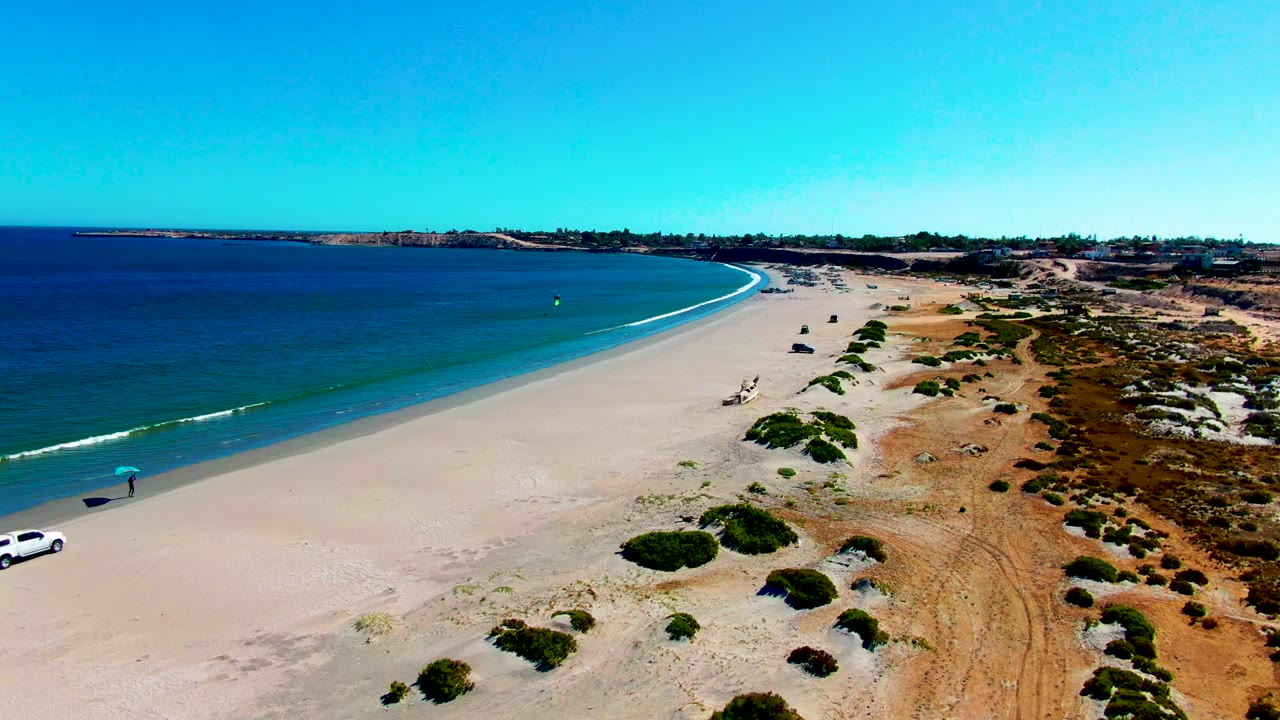 Aerial view of San Juanico beach in Baja California Sur with blue ocean, pale sand, dune scrub, a white car at the lower left, and a person under an umbrella near the shore.