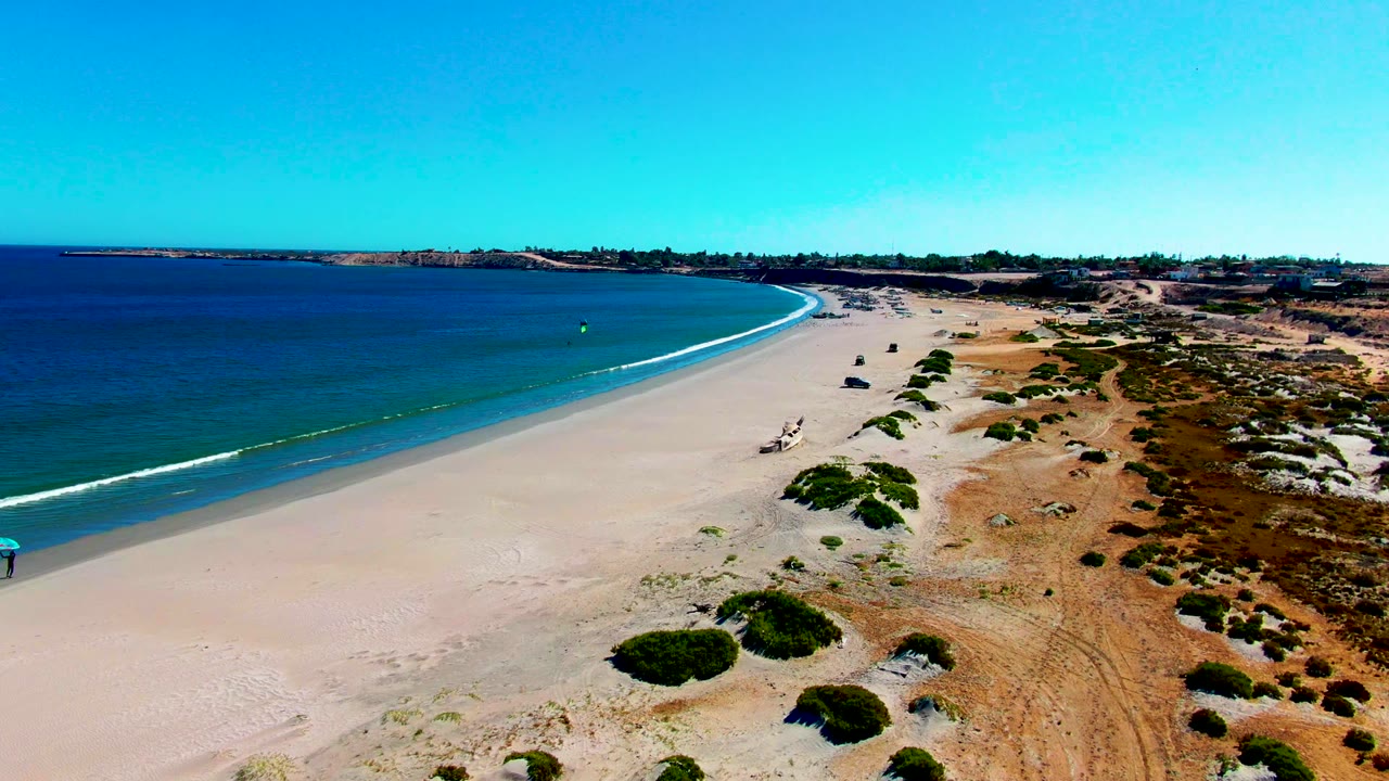 Aerial beach view with ocean, surf line, dunes, and no visible people.