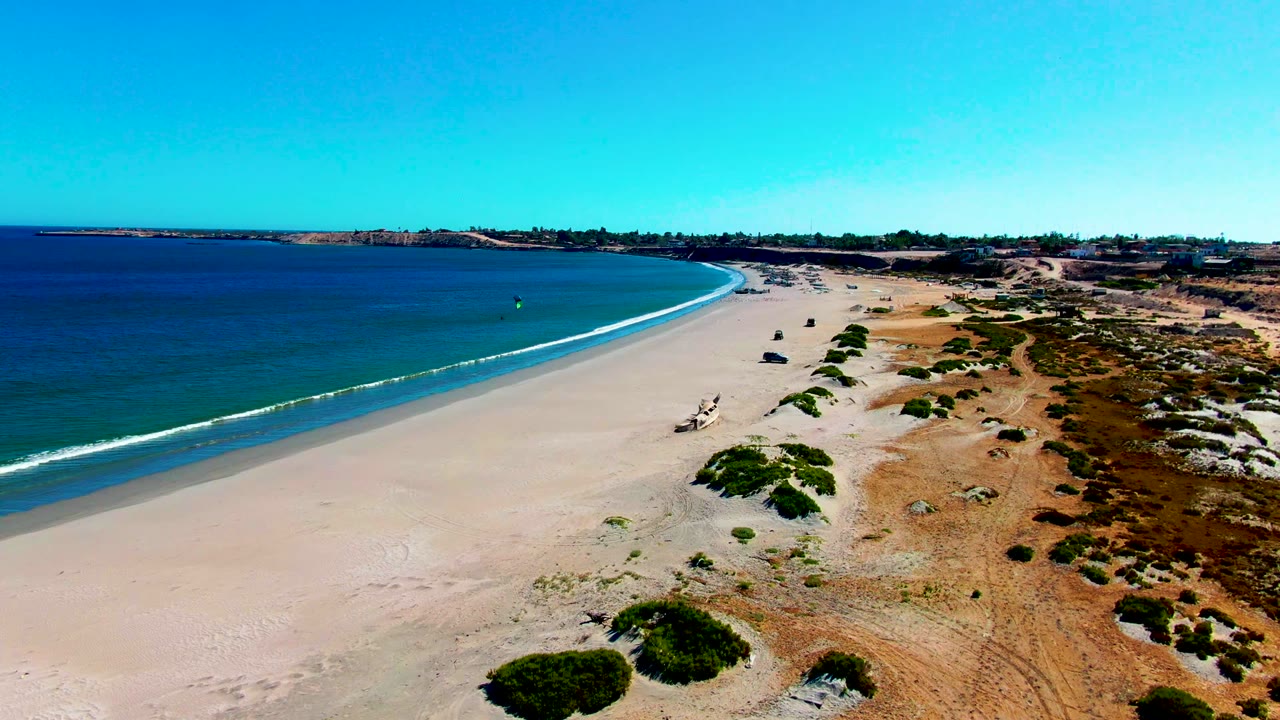 Aerial San Juanico coastline with blue water on the left, pale open sand in the center, dune scrub on the right, and no visible people.