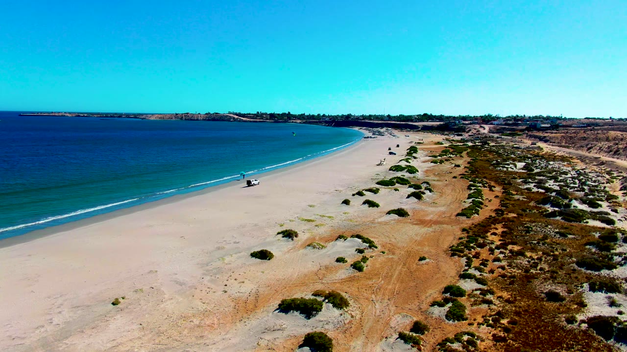 Aerial view in San Juanico, Baja California Sur, Mexico, with a white 4x4 on pale sand near the shoreline, blue water on the left, dune scrub on the right, and no clearly recognizable people.