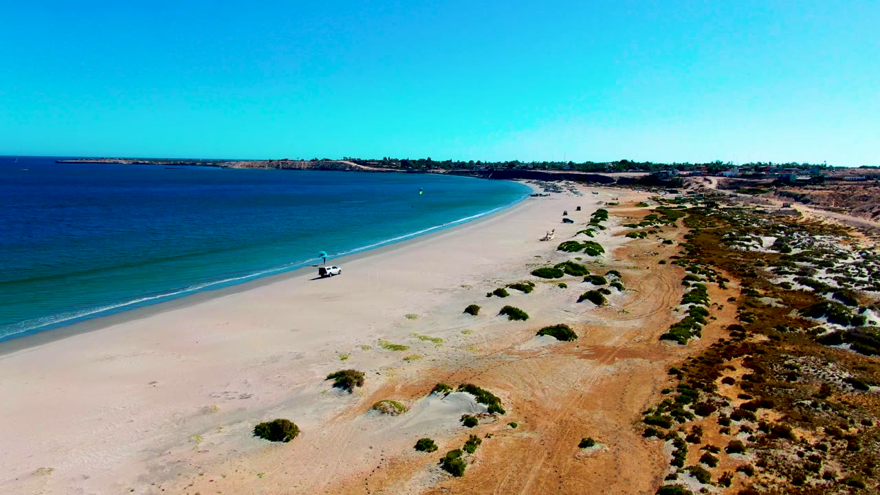 High drone view over San Juanico beach with the white 4x4 near the waterline, a small beach user or object beside it, low surf, dry sand, shrubs, and no recognizable faces.