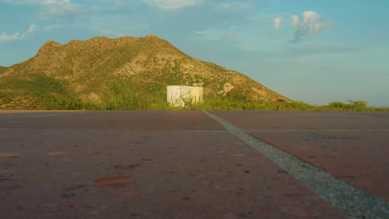 Opening frame of a low paved road view in Cabo Pulmo, Baja California Sur, with a diagonal white line, green scrub, a small white structure, a sunlit mountain, blue sky, and no visible people.