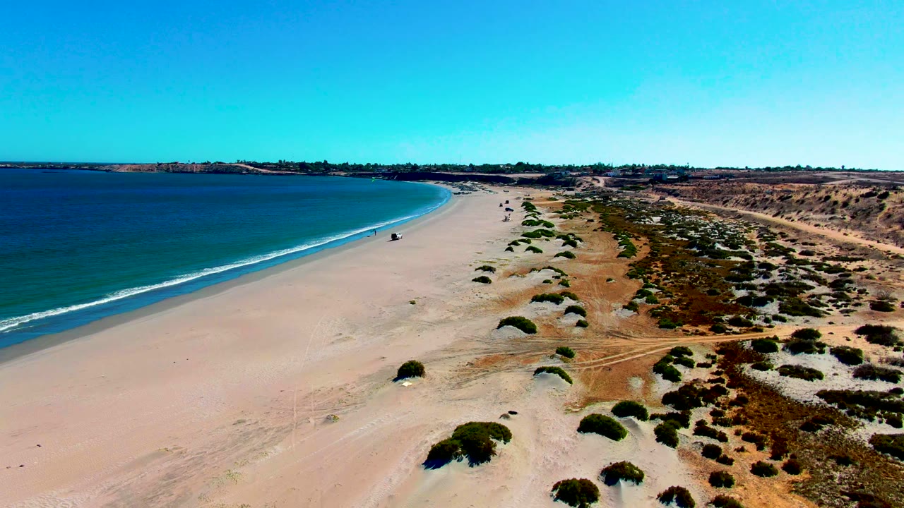 Aerial frame over San Juanico, Baja California Sur, with blue ocean on the left, a wide sandy beach curving ahead, dune shrubs on the right, and a tiny person near the shoreline.