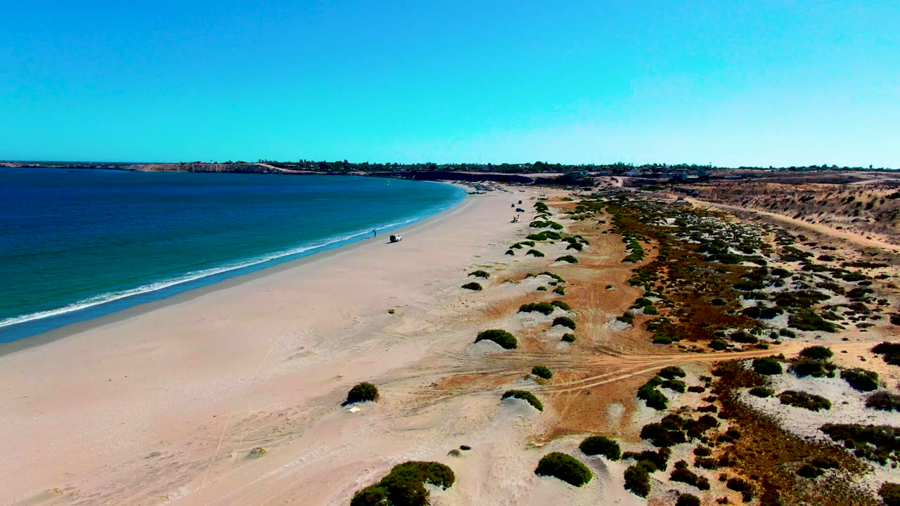 Wide aerial beach view with ocean, pale sand, dunes, shrubs, and a small walker.