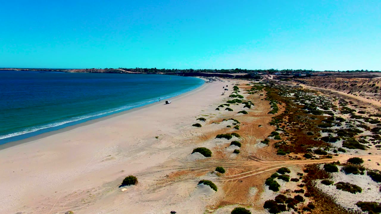 San Juanico aerial beach frame with a curved strip of blue water, white wave edge, open sand, scattered dune shrubs, dry tracks near the dunes, and a distant person close to the surf.