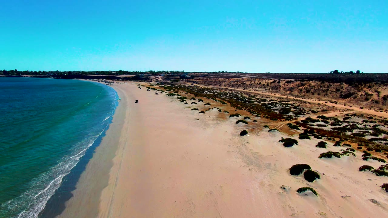 Opening aerial view over San Juanico, Baja California Sur, with blue ocean at left, pale sand beach in the center, low vegetated dunes at right, and no visible people.