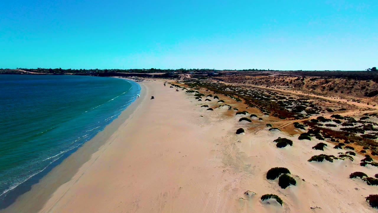 Wide drone frame over San Juanico beach, showing a smooth sand corridor between small rolling waves on the left and scattered dune vegetation on the right, with no visible people.