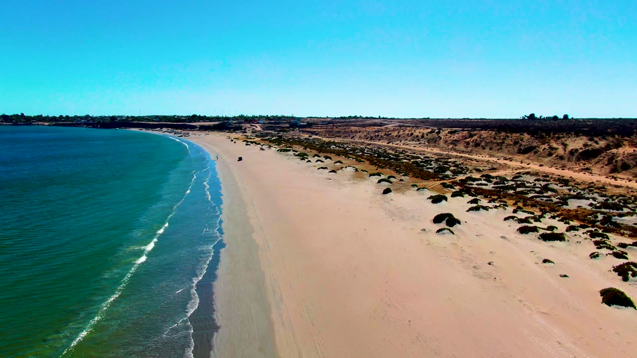 Aerial view over San Juanico, Baja California Sur, with turquoise water on the left, a pale sandy beach through the center, low dunes on the right, and no visible people.