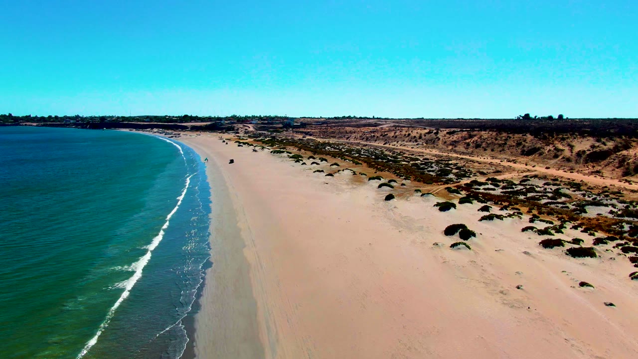 Curving shoreline with small white waves and sparse vegetation beyond the sand.