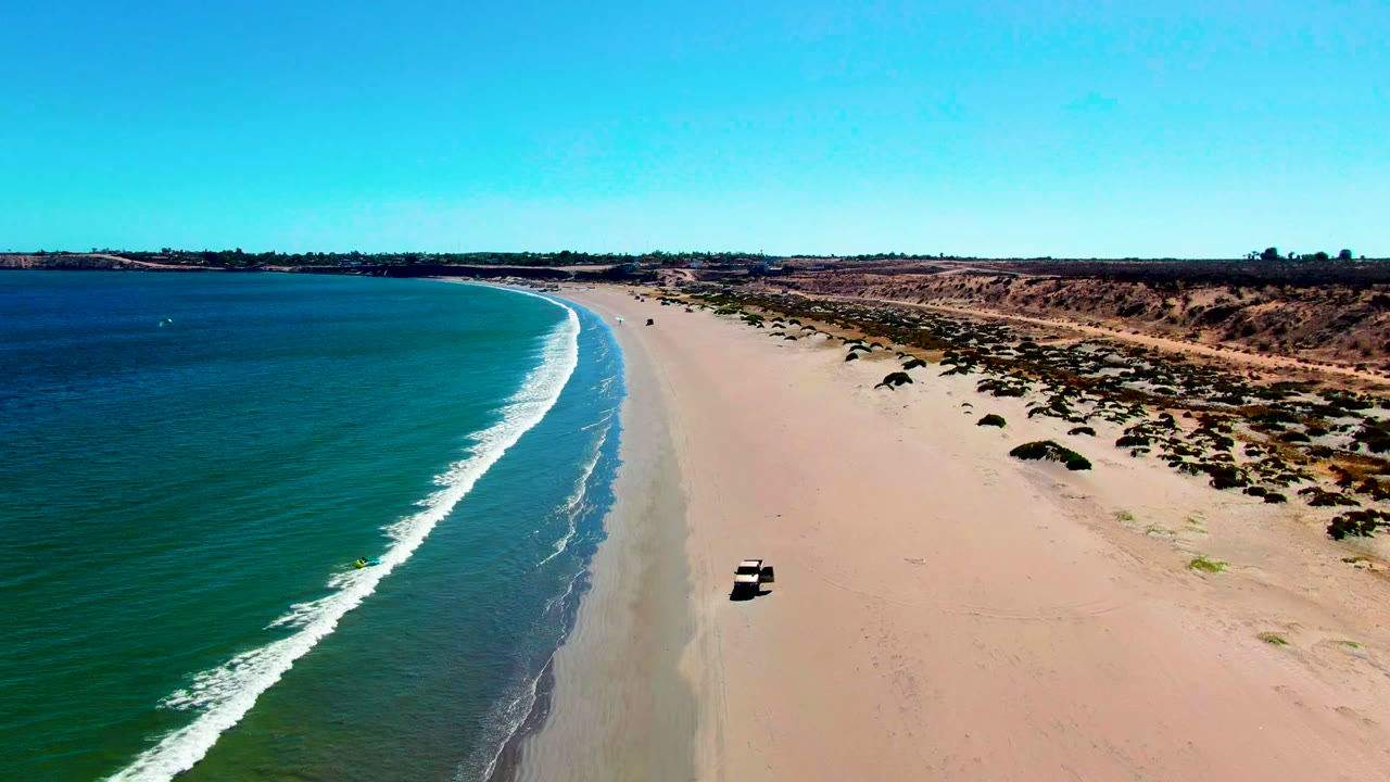 Aerial view in San Juanico, Baja California Sur, with a light vehicle on open sand near the waterline, blue ocean to the left, dunes to the right, and no visible people.