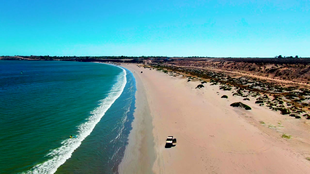 Vehicle driving along a wide beach with waves curving onto shore.