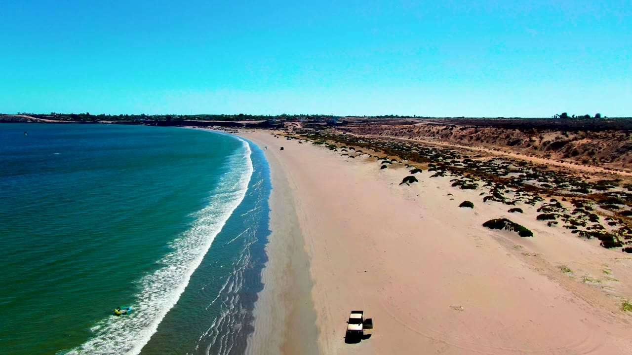 High aerial frame in San Juanico showing the vehicle closer to the lower right edge, green-blue water and foam on the left, open sand through the center, and no visible people.