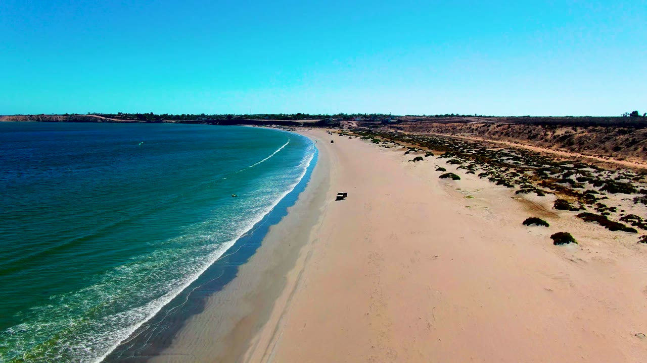 Aerial frame over San Juanico, Baja California Sur, showing blue ocean at left, a broad sandy beach, low scrubland to the right, a small vehicle on the shore, and tiny distant beachgoers visible.