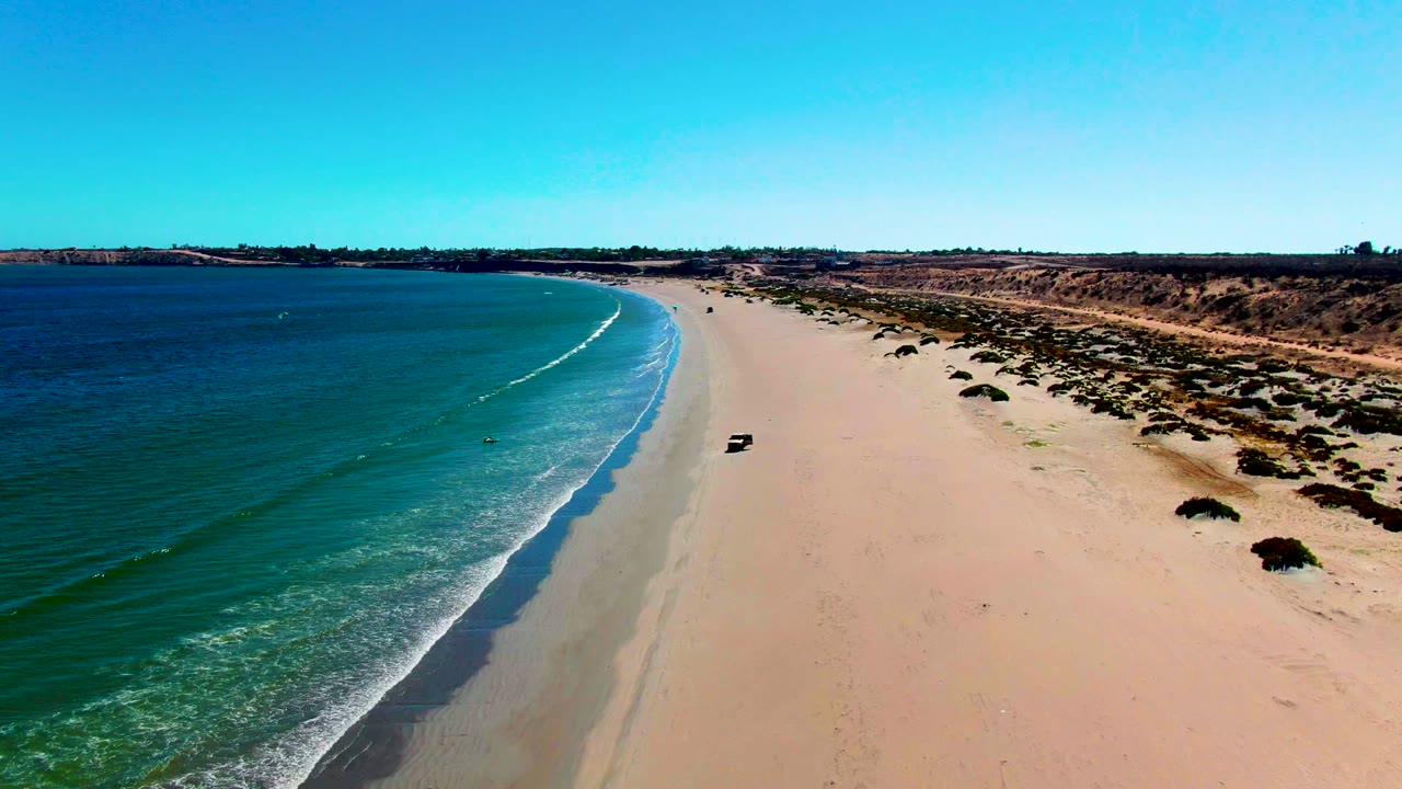 Drone view of sandy coastline with a vehicle near the waterline.