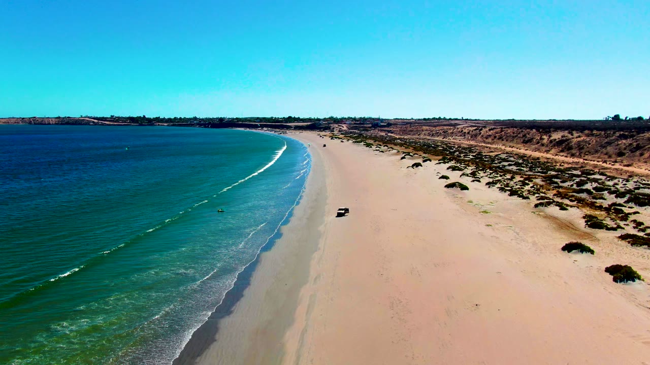 High oblique aerial frame of the San Juanico shoreline, with darker blue water at left, a narrow white wave approaching shore, a small vehicle on the pale sand, scrub-covered beach edge, and distant people visible as tiny figures.