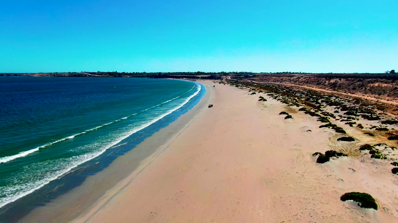 Aerial frame over San Juanico, Baja California Sur showing a small dark car on a wide sandy beach beside teal water and white surf, with no visible people.