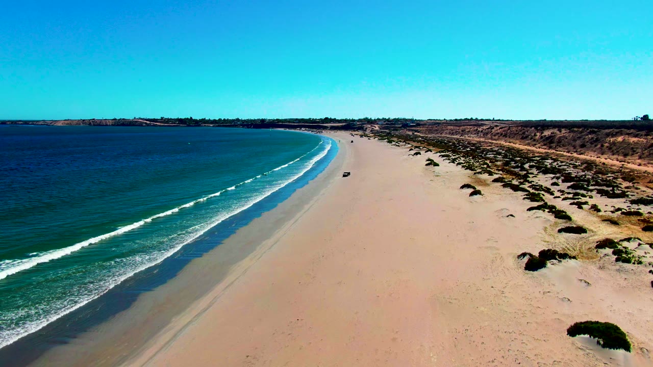 Car moving along a broad beach with waves on the left