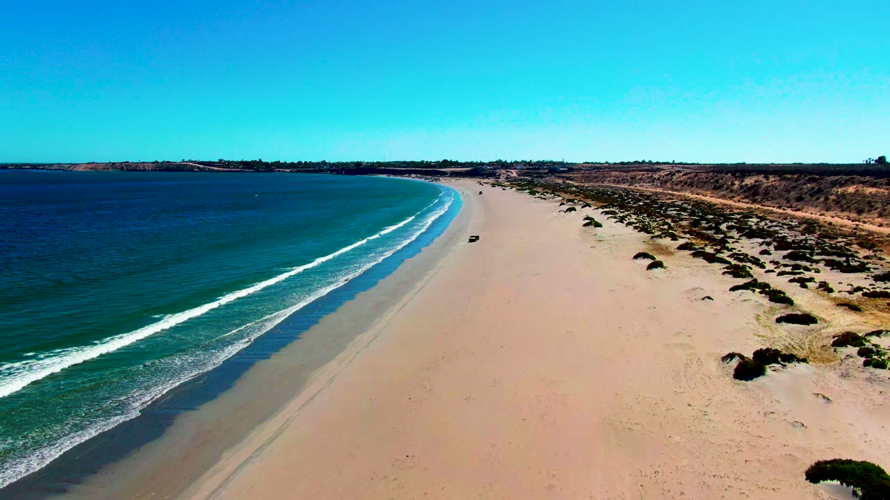 High drone perspective over San Juanico beach showing a small dark car near the center sand, green-blue ocean on the left, dune vegetation on the right, and no visible people.