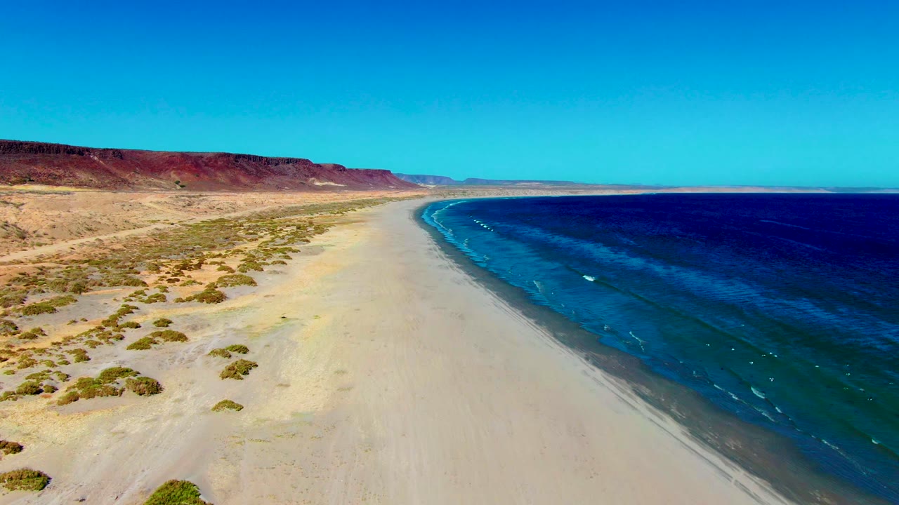 Aerial view of San Juanico, Baja California Sur coastline with pale sand on the left, deep blue water on the right, reddish desert bluffs in the distance, and no visible people.