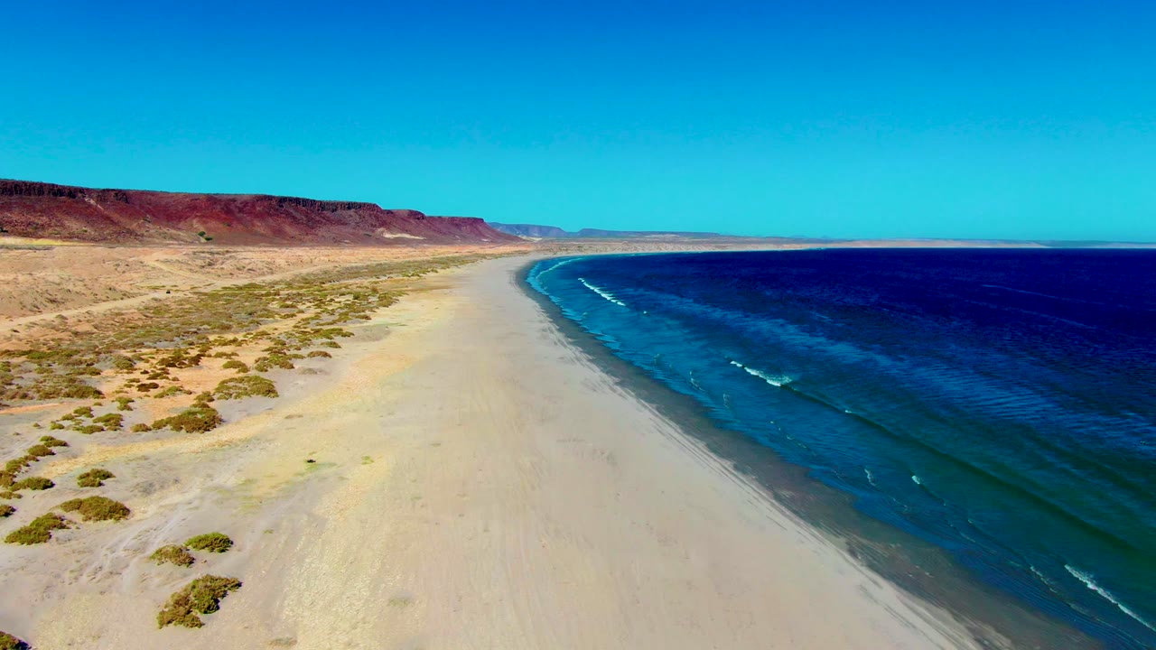 Aerial view along the San Juanico shoreline as the beach curve becomes more pronounced, with ocean whitecaps closer to frame center and no visible people.
