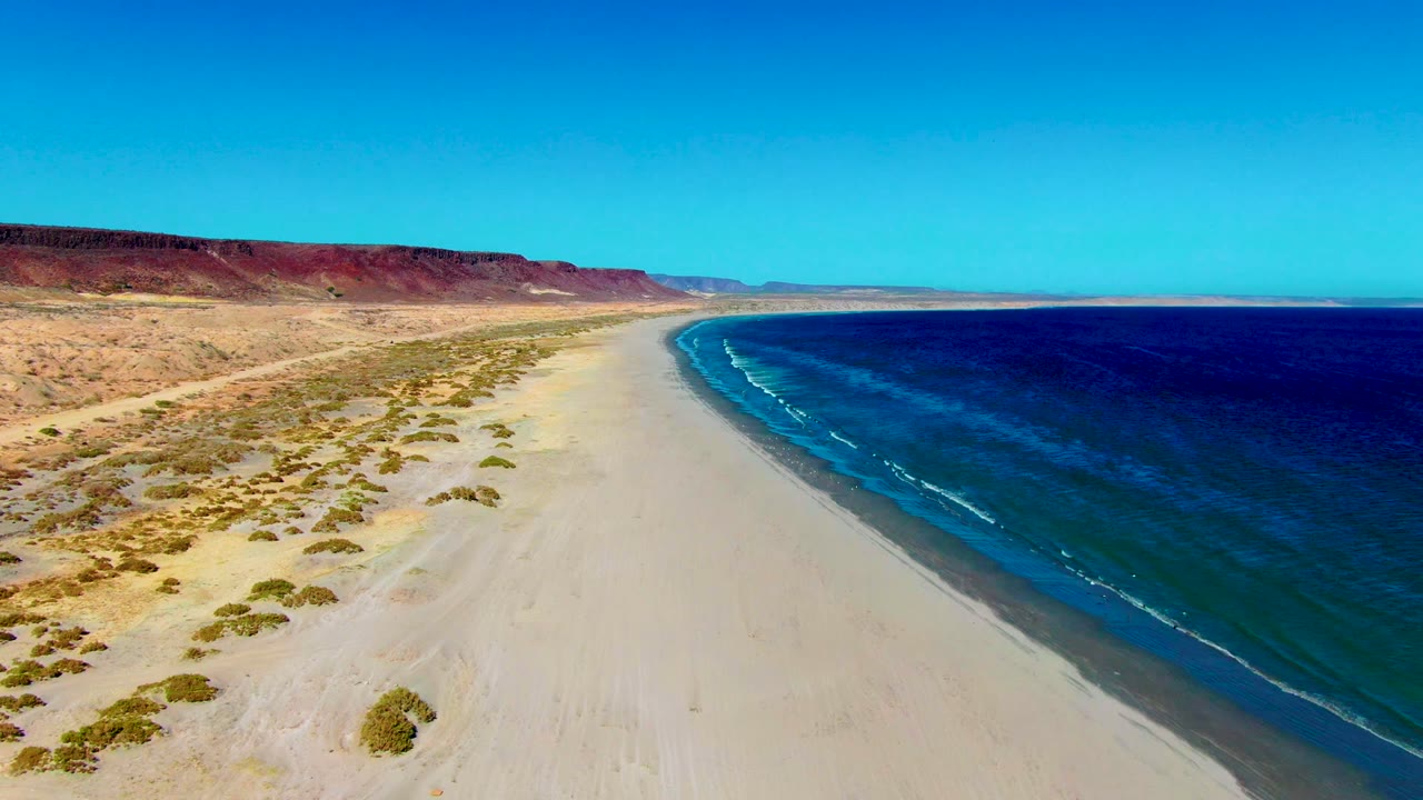 Aerial view over San Juanico beach with desert scrub and dunes on the left, blue ocean on the right, reddish cliffs in the distance, and no visible people.