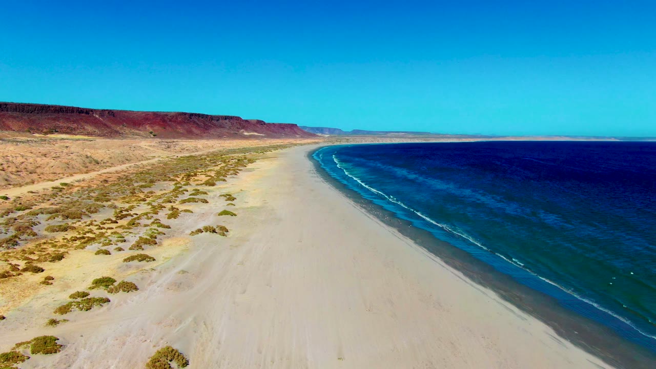 Wide aerial of empty sand beach with surf line and arid coastline.