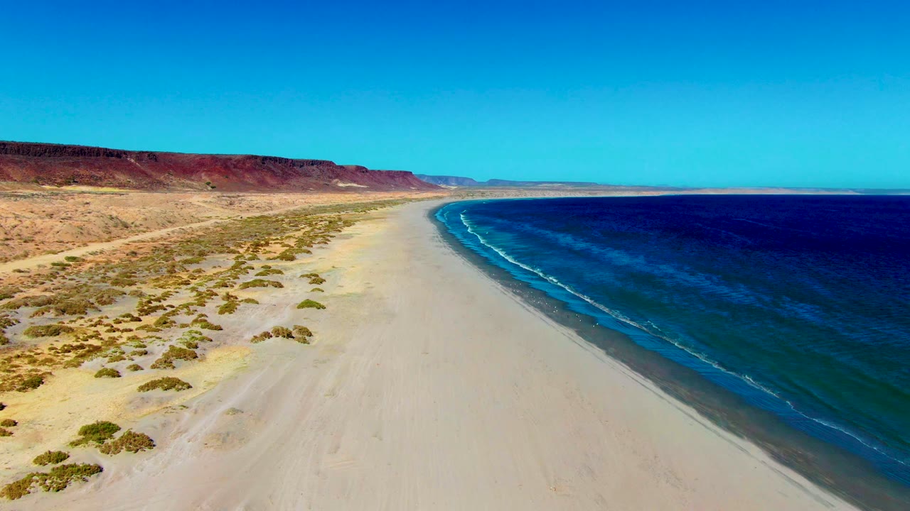 Aerial frame of the San Juanico beach curve with ocean water occupying the right half, dry scrubland on the left, a clear sky, and no visible people.