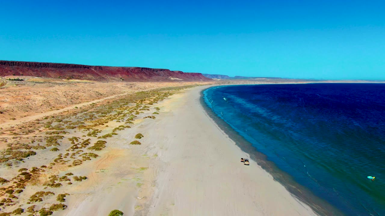 Aerial frame from San Juanico, Baja California Sur showing a small vehicle on pale sand near the waterline, blue ocean to the right, desert scrub and red cliffs to the left, and no visible people.