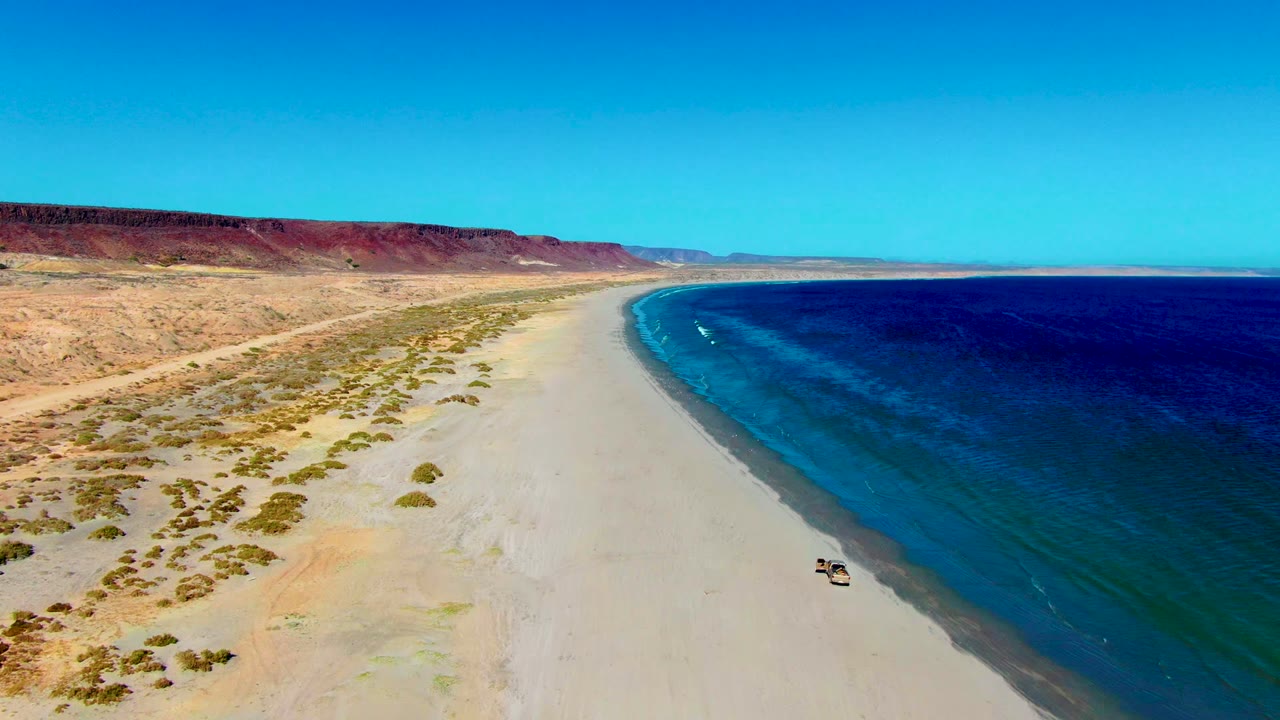 Wide aerial shoreline with a vehicle driving along the beach near the water.