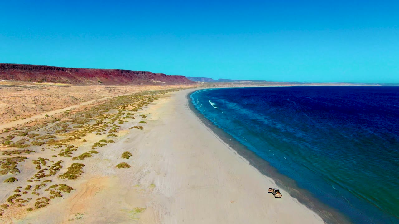 Aerial view over San Juanico beach with the vehicle near the lower right sand, a long arc of blue coastline, dry scrubland, reddish cliffs, and no visible people.