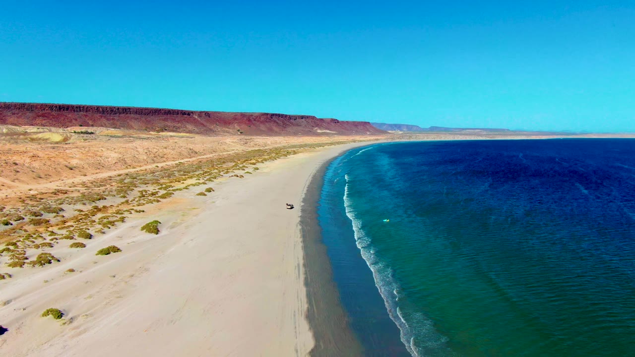 Aerial view in San Juanico, Baja California Sur with a small vehicle on pale sand, clear blue ocean to the right, desert scrub inland, distant bluffs, and no visible people.
