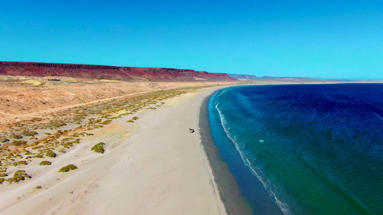 Wide aerial frame over San Juanico with the vehicle near the wet sand edge, turquoise shallows, darker offshore water, scrub-covered dunes, red-brown bluffs, and no visible people.