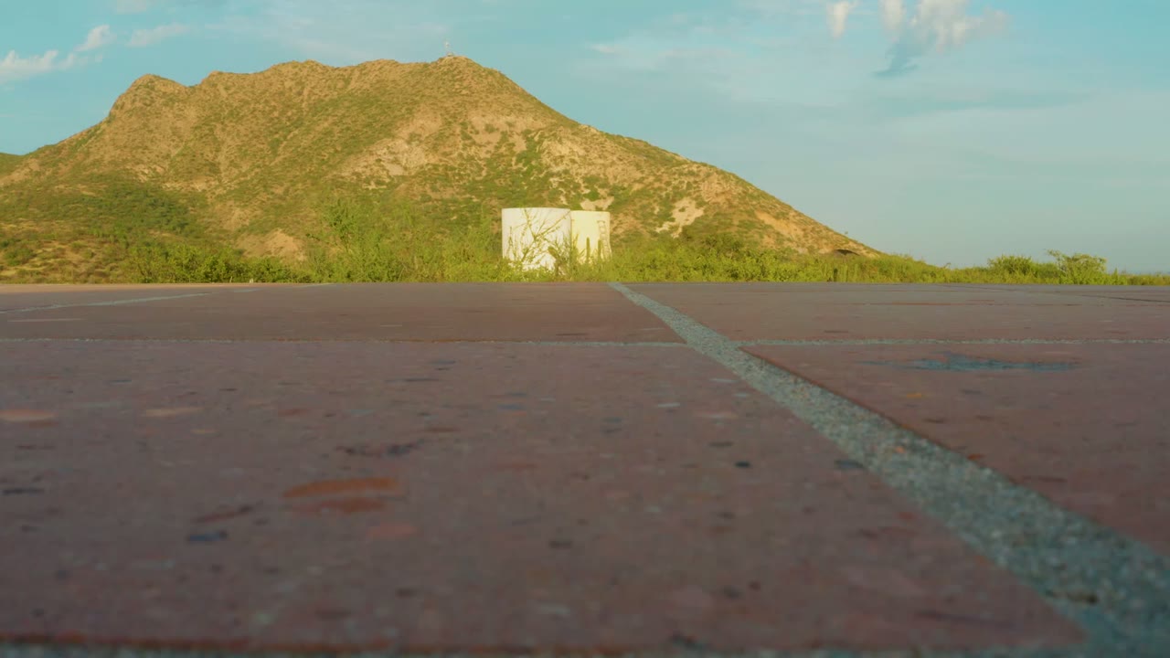 At 0:00, a low view in Cabo Pulmo, Baja California Sur, Mexico shows reddish asphalt, a pale road line, scrub greenery, a small white structure, a brown-green mountain, blue sky, and no visible people.